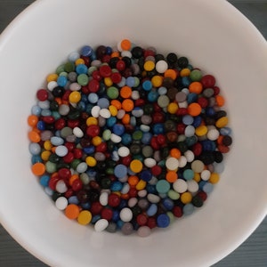 May include: A white bowl filled with a colorful assortment of small, round glass beads. The beads are in various colors, including red, orange, yellow, green, blue, and white. The bowl is set against a blue-green surface.