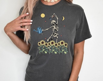 Pode incluir: Uma camiseta cinza escura com um gráfico de um esqueleto regando girassóis. O esqueleto está segurando um regador e uma ferramenta de jardinagem. O gráfico também inclui uma lua, um sol e estrelas.