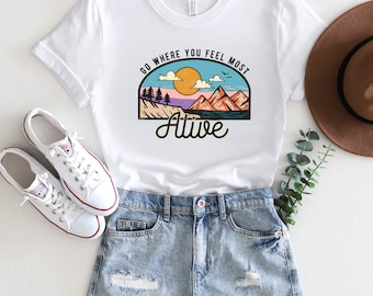 Puede incluir: Camiseta blanca con un gráfico de un paisaje de montaña con un sol y el texto "Go where you feel most alive".