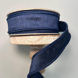 May include: A spool of navy blue ribbon with a satin finish. The ribbon is wrapped around a wooden spool and is displayed on a white pedestal.