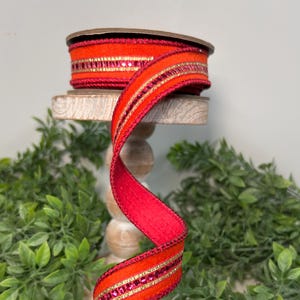 May include: Red velvet ribbon with a gold and red trim and sparkly pink accents. The ribbon is curled on a wooden stand.