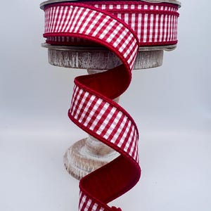 May include: A spool of red and white gingham ribbon, displayed on a wooden stand. The ribbon is a vibrant red with a white checkered pattern. The ribbon is spiraling down from the spool, showcasing its texture and design. Perfect for crafting and gift wrapping.