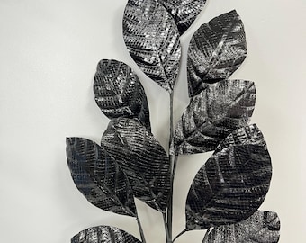 May include: A black artificial plant stem with multiple large, textured leaves. The leaves have a reptile-like pattern and a metallic sheen. The stem is dark and the leaves are arranged in an upward direction. The plant is against a white background.