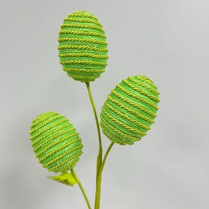 May include: A green stem with three green, sparkly, egg-shaped ornaments. The ornaments are made of a woven material and have a spiral pattern.