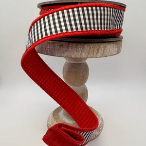 May include: A spool of decorative ribbon with a black and white gingham pattern and a red border. The ribbon is displayed on a wooden stand, with a portion of the red ribbon unfurled. This ribbon is suitable for crafts and gift wrapping.