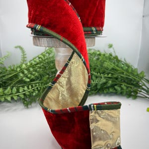 May include: A roll of wired ribbon featuring a deep red velvet texture on one side and a metallic gold finish on the reverse. The edges are trimmed with a dark green, red, and white plaid pattern, providing a structured border to the fabric.