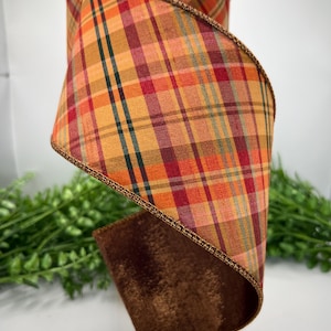 May include: A spool of wired ribbon featuring a plaid pattern in shades of orange, red, yellow, and dark green. The ribbon has a metallic copper-colored edge and a solid dark brown velvet-textured underside, coiled to display both sides.