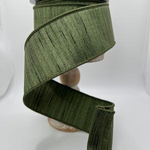 May include: A spool of olive green ribbon with a textured, vertical ribbed design. The ribbon is displayed on a wooden spool holder. The ribbon is a wide, decorative trim, suitable for crafts and gift wrapping.