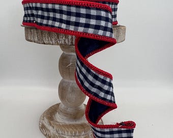 May include: A spool of navy blue and white gingham ribbon with a red stitched edge. The ribbon is displayed on a decorative wooden stand, with a portion of the ribbon unrolled, showcasing its pattern and texture.