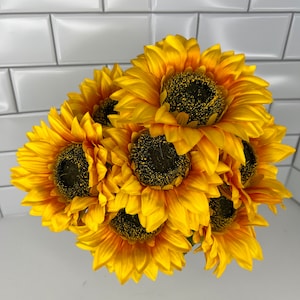 May include: A bouquet of seven artificial sunflowers with yellow petals and dark brown centers. The sunflowers are arranged in a cluster, with the petals overlapping.