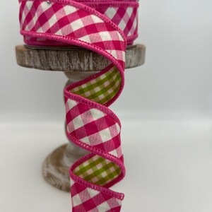May include: A roll of decorative ribbon with a pink and white gingham pattern on one side and a green and white gingham pattern on the other. The ribbon is displayed on a wooden stand.
