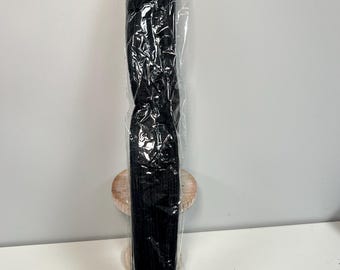 May include: A long, black, packaged item, possibly hair extensions or a similar product, is displayed vertically. The item is encased in clear plastic, revealing its dark color and texture. The background is a plain white surface.
