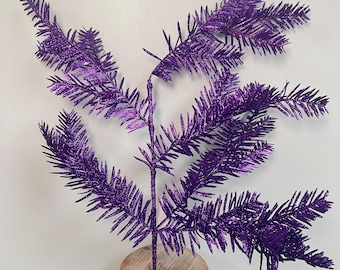 May include: A single, artificial purple glittered pine branch. The branch is displayed on a small, round, wooden pedestal. The pine needles are a deep purple color and covered in glitter.