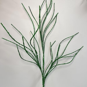 May include: A decorative green glittered branch with multiple slender stems. The branch is set on a small, round wooden base. The stems are a vibrant green color and are covered in glitter.