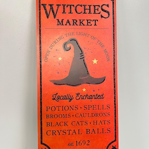 May include: A rectangular orange sign with black glitter trim and text that reads "Witches Market". The sign features a witch's hat, stars, and a broom graphic. Additional text includes "Locally Enchanted" and a list of items.