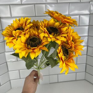 May include: A bouquet of seven artificial sunflowers with yellow petals and brown centers. The sunflowers are arranged in a bunch and have green stems.