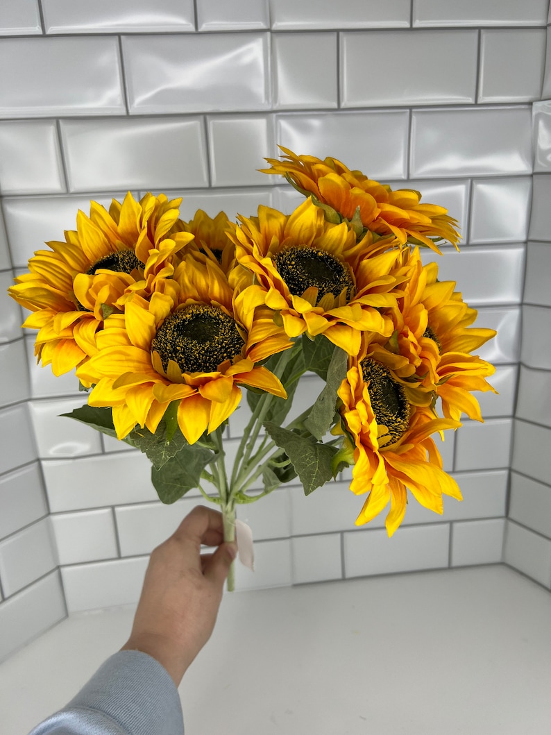 May include: A bouquet of seven artificial sunflowers with yellow petals and brown centers. The sunflowers are arranged in a bunch and have green stems.