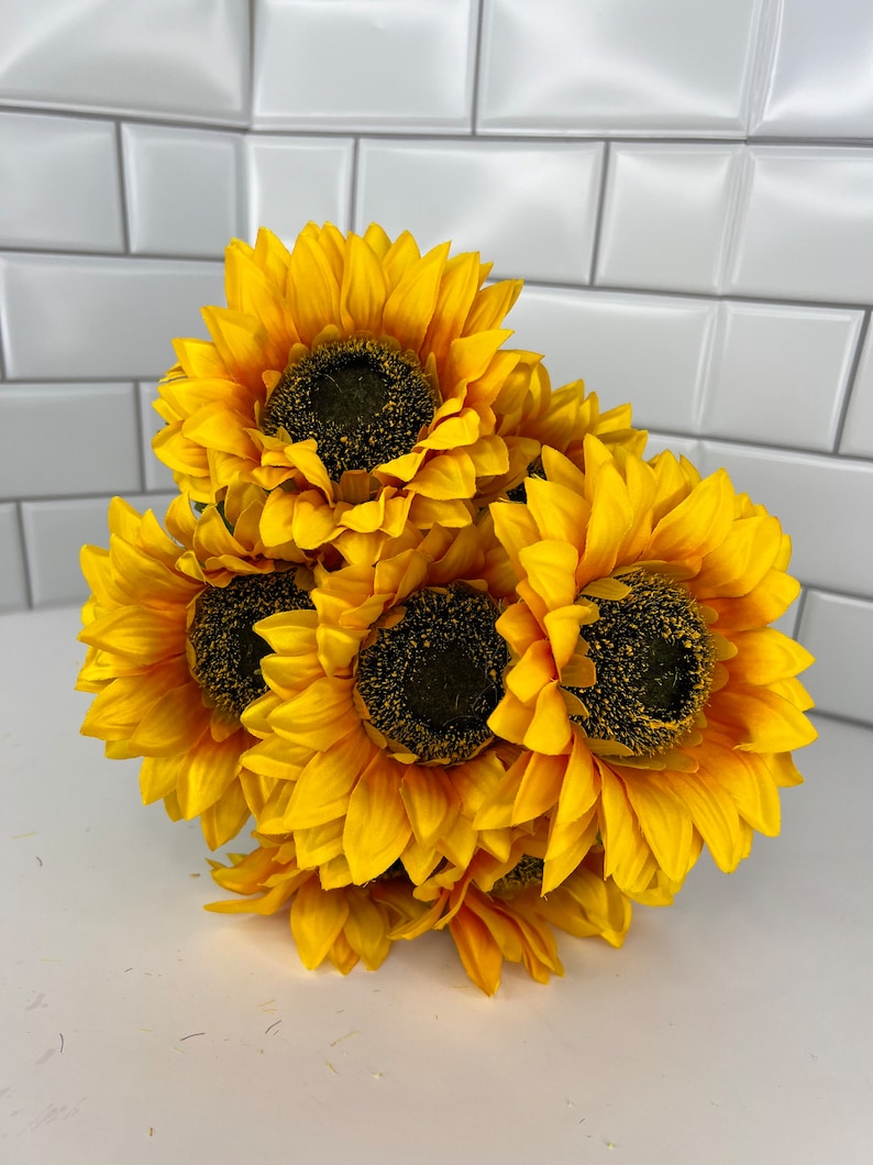 May include: A bouquet of six artificial sunflowers with yellow petals and dark brown centers. The sunflowers are arranged in a cluster, with the petals overlapping.