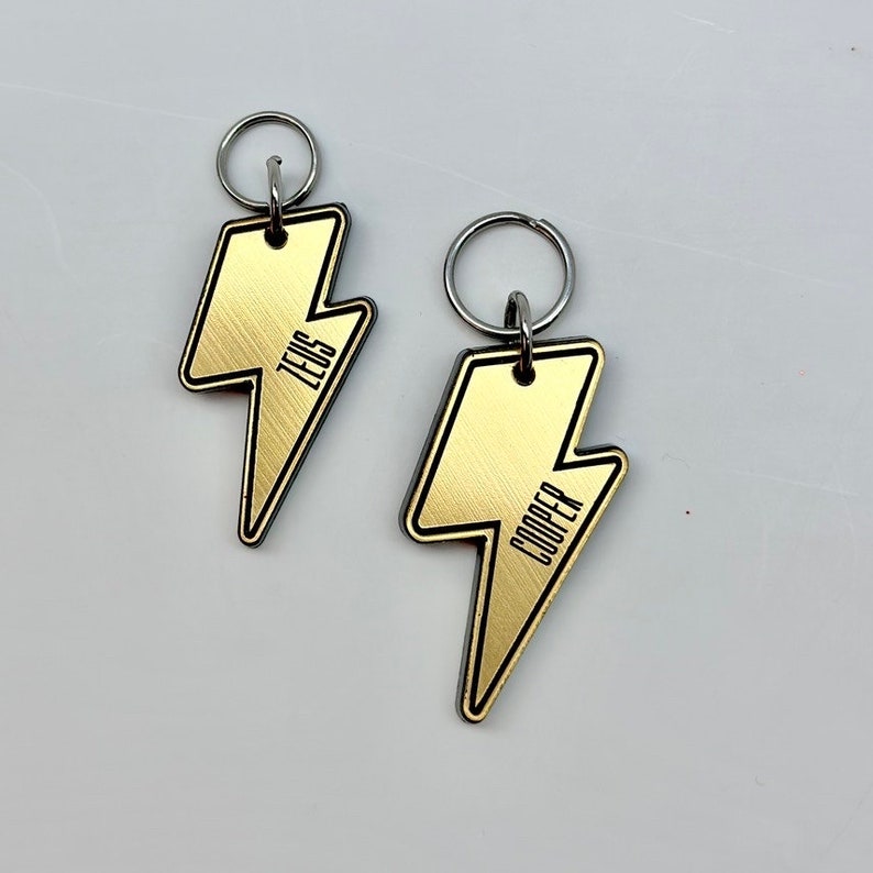 May include: Two gold lightning bolt shaped keychains with black outlines. The keychains have the names "Zeus" and "Cooper" engraved on them.