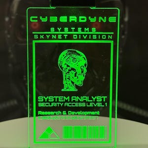 Könnte beinhalten: Ein grüner Plastik-Ausweis mit dem Text "CYBERDYNE SYSTEMS SKYNET DIVISION" und einer Grafik eines Roboterkopfes. Der Text "SYSTEM ANALYST SECURITY ACCESS LEVEL 1" und "Research & Development 10#2605199508291997" ist ebenfalls enthalten.