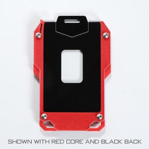 May include: A rectangular metal card holder featuring a vibrant red frame and a smooth black center panel. The design includes a central rectangular cutout, four silver-toned screws at the corners, and a top slot for attachment. Text reads: SHOWN WITH RED CORE AND BLACK BACK.