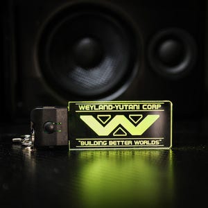 Könnte beinhalten: Ein schwarzer und grüner Schlüsselanhänger mit dem Weyland-Yutani Corp. Logo und dem Text "Building Better Worlds".