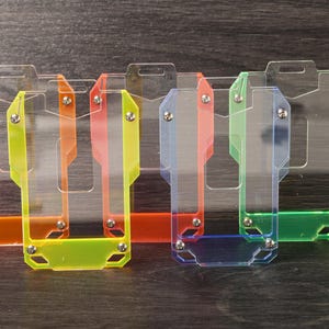 May include: A set of five rectangular badge holders made of transparent acrylic with colored borders in orange, yellow, red, blue, and green. Each holder features a top slot for a lanyard and small metal screws securing the clear front panel.