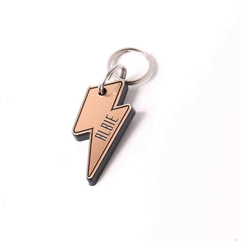 May include: A gold lightning bolt shaped dog tag with a black border and the name "Albie" engraved on it. The tag is attached to a silver key ring.