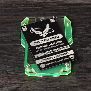 May include: A black rectangular identification card labeled AREA 51 FULL ACCESS CHRIS JONES EXTRATERRESTRIAL AFFAIRS PRIORITY PERSONNEL BIOMETRIC AUTHENTICATION held within a neon green translucent acrylic frame secured by four metal screws.
