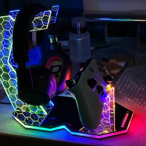 Peut inclure: Un casque de jeu noir repose sur un support hexagonal éclairé par des LED multicolores. Une manette de jeu blanche est également sur le support. Le support a une base noire et un design en nid d'abeille. Le support est illuminé de lumières rouges, bleues et vertes.