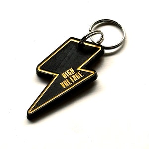 May include: Black lightning bolt keychain with a gold inscription that reads "HIGH VOLTAGE".