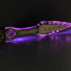 May include: A black and clear plastic tool with a purple glow. The tool has a handle with the word "QUITO" printed on it and a pointed tip.