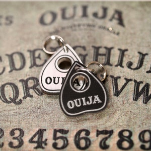Pode incluir: Dois pingentes de planchette Ouija prateados e pretos com um furo para uma corrente ou cordão. Os pingentes estão em um tabuleiro Ouija vintage com letras e números pretos.