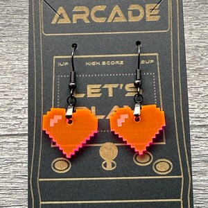 May include: A pair of orange pixel heart earrings with pink outlines hanging from black hooks. The earrings are displayed on a black card with the word "ARCADE" printed in gold letters.