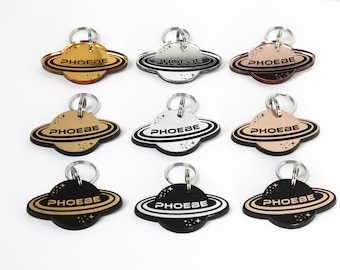 May include: Nine keychains featuring a gold, silver, bronze, or black planet design with a gold ring around it. The planet has a black inscription that reads "PHOEBE" and small white stars.