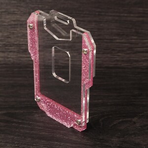 May include: A clear acrylic badge holder featuring a pink glitter border. The rectangular frame is constructed from two layers of transparent material held together by four small silver-toned metal screws, with a slot at the top for a lanyard.