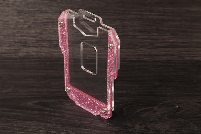 May include: A clear acrylic badge holder featuring a pink glitter border. The rectangular frame is constructed from two layers of transparent material held together by four small silver-toned metal screws, with a slot at the top for a lanyard.
