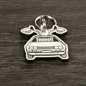 Puede incluir: Un llavero plateado con forma de coche DeLorean con el nombre "Emmett" impreso en el parabrisas. El llavero tiene un borde negro y un llavero plateado. Las puertas de ala de gaviota del coche están abiertas.