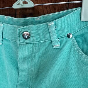 May include: Close-up of turquoise denim shorts. The shorts feature a button closure, belt loops, and white stitching. The metal button has a logo. The shorts are hanging on a white hanger against a wooden background.
