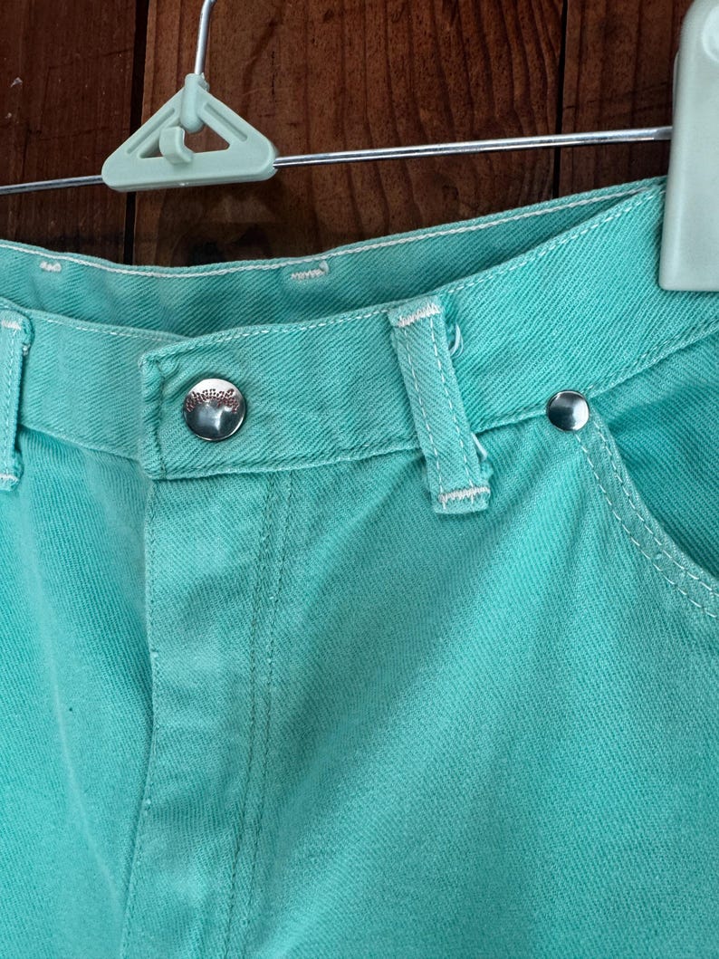 May include: Close-up of turquoise denim shorts. The shorts feature a button closure, belt loops, and white stitching. The metal button has a logo. The shorts are hanging on a white hanger against a wooden background.