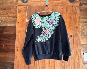 May include: Black sweatshirt with a floral design painted on the front. The flowers are pink, purple, and green with gold accents.