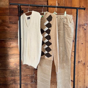May include: A clothing rack displays a cream-colored V-neck sweater vest, a tan argyle sweater, and tan corduroy pants. The garments hang against a wooden backdrop, showcasing a classic, preppy style.