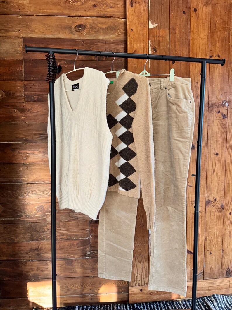 May include: A clothing rack displays a cream-colored V-neck sweater vest, a tan argyle sweater, and tan corduroy pants. The garments hang against a wooden backdrop, showcasing a classic, preppy style.