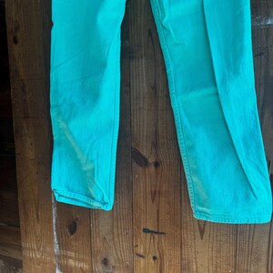 May include: A pair of turquoise-colored pants. The pants are made of a solid fabric and have a classic five-pocket design. The pants are displayed against a wooden background.