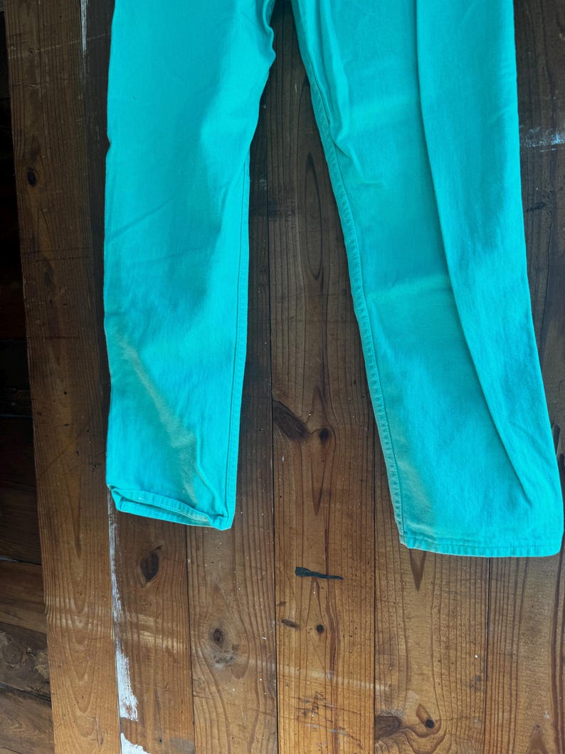 May include: A pair of turquoise-colored pants. The pants are made of a solid fabric and have a classic five-pocket design. The pants are displayed against a wooden background.