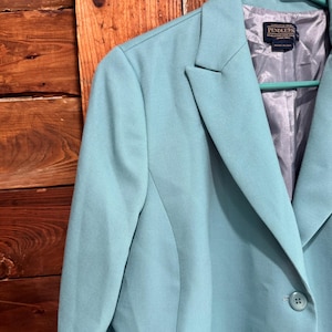 May include: A light blue blazer with a two-button closure and notched lapels. The blazer is hanging on a green hanger against a wooden background. The interior lining is a light gray color. The brand label is visible.
