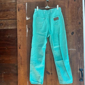 May include: A pair of turquoise Wrangler jeans hanging on a hanger. The jeans have a high waist, classic five-pocket design, and a brown leather patch on the back. The denim has a faded, vintage look.
