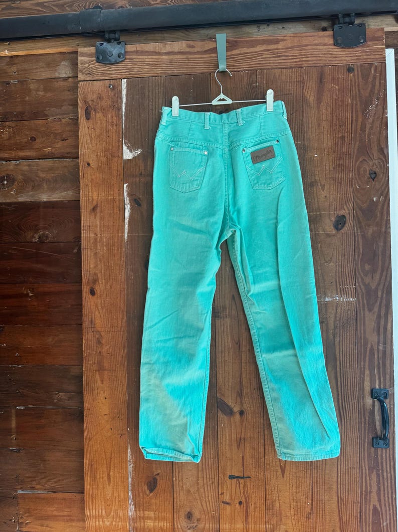 May include: A pair of turquoise Wrangler jeans hanging on a hanger. The jeans have a high waist, classic five-pocket design, and a brown leather patch on the back. The denim has a faded, vintage look.
