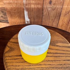 May include: A yellow and white thermos food container with the word "THERMOS" printed on the lid.