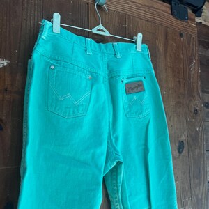 May include: Turquoise Wrangler jeans hanging on a white hanger. The jeans have a classic five-pocket design with white stitching and a brown leather patch with the Wrangler logo on the back pocket. The jeans are made of a sturdy denim material.